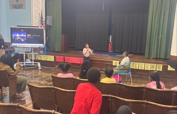  Student speaking into microphone in front of auditorium while Ms. Lee sits next to her in chair.